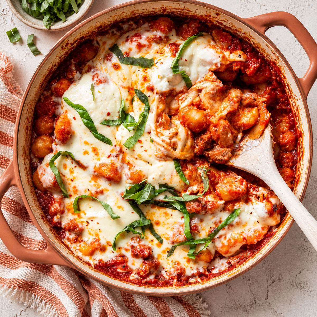 Overhead shot of a baked pasta dish in a terracotta pot, topped with melted cheese, basil leaves, and a wooden spoon serving food. A striped cloth napkin and a small bowl of chopped herbs are visible on a light stone surface.