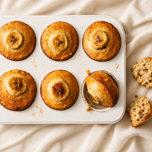 Six banana muffins in a white six-cup muffin tin on a cream fabric background, with two muffins broken open to show the crumb. The muffins have a golden-brown baked surface with a sliced banana visible in the center of each.