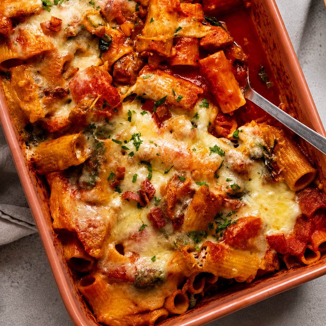 A close-up overhead shot of a chicken and bacon pasta bake in a rectangular terracotta dish. The pasta is mixed with red tomato sauce, melted cheese, herbs, and small pieces of meat, with a metal serving spoon resting on the right side.