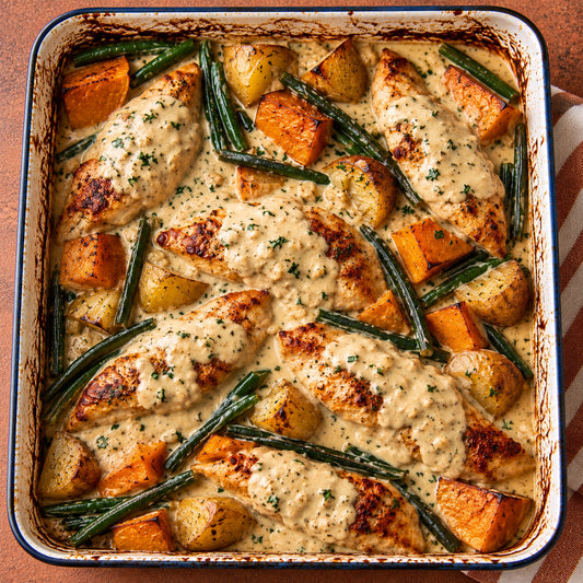 Overhead view of a baked chicken and vegetable casserole in a square baking dish. The dish features pieces of chicken covered in a creamy beige sauce, mixed with roasted orange sweet potatoes, yellow potatoes, green beans, and chopped green herbs, all in a browned ceramic dish.