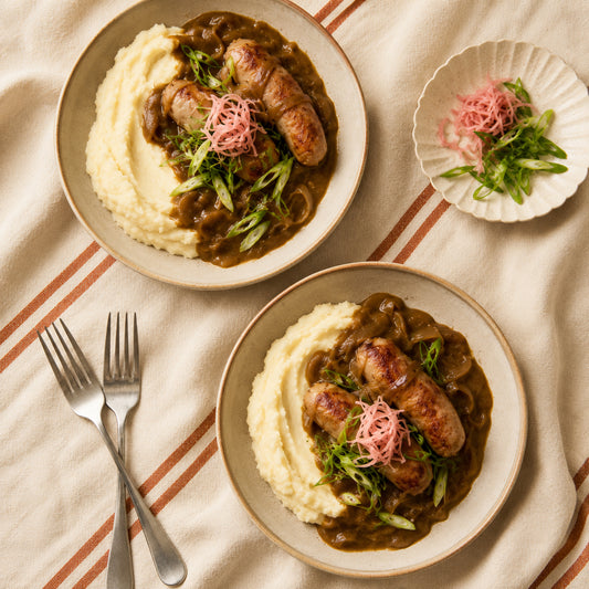 Two bowls of sausage and mashed potatoes in japanese style curry sauce garnished with green onions and pink shredded pickled onions, arranged on a striped beige cloth with two forks and a small side plate.
