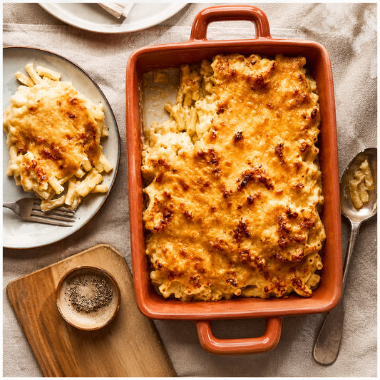 A baked macaroni and cheese dish in a terracotta-colored rectangular casserole dish, styled on a beige fabric surface with a plated serving, fork, spoon, cutting board, and small bowl of pepper.