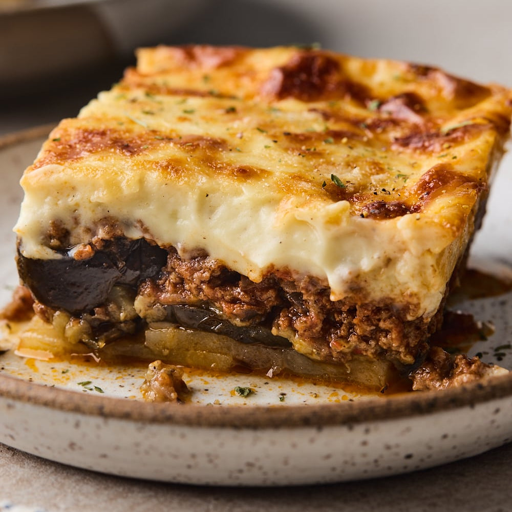 A close-up of a baked square slice of Moussaka on a speckled ceramic plate, showing layers of melted cheese, meat sauce, eggplant, and pasta against a warm neutral background.