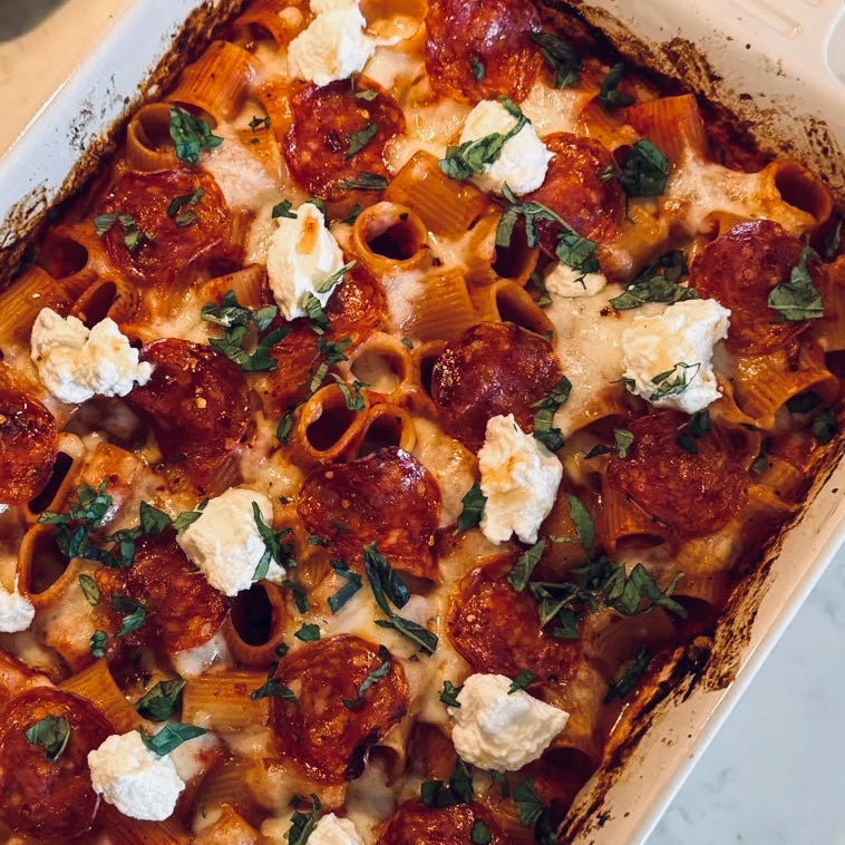 A close-up overhead view of a baked pasta casserole in a white ceramic baking dish, topped with pepperoni, melted cheese, and dollops of white cream with chopped green herbs.