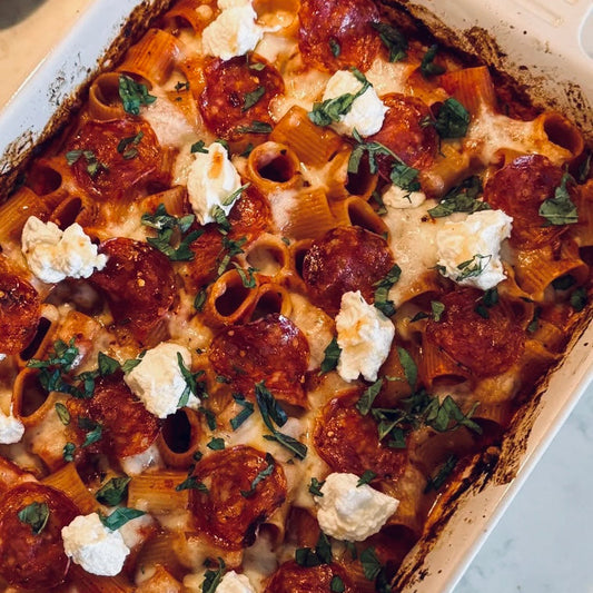 A close-up overhead view of a baked pasta casserole in a white ceramic baking dish, topped with pepperoni, melted cheese, and dollops of white cream with chopped green herbs.