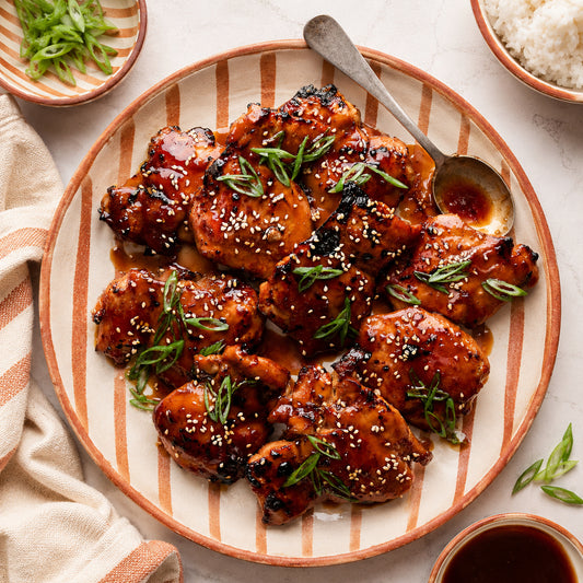 Glazed teriyaki chicken pieces on a striped ceramic plate with a spoon, surrounded by bowls of rice, green onions, and sauce in a styled food setting.