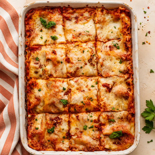 A baked dish of square-cut vegetable lasagna in a white ceramic baking pan, topped with melted cheese, red sauce, and fresh green herbs, photographed from above on a light beige surface with a striped cloth at the left edge.
